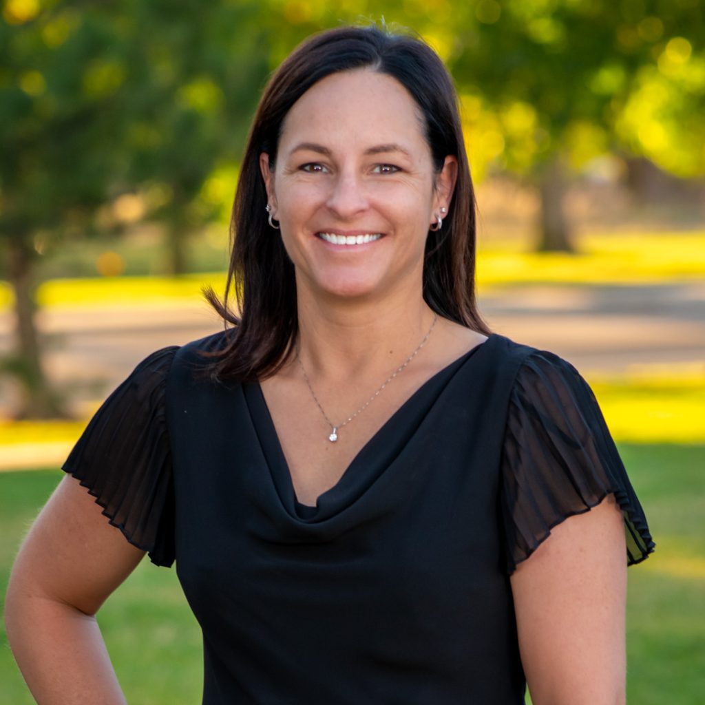 A woman with dark hair, wearing a black blouse and a necklace, smiles while standing outdoors in a park with green grass and trees, embodying the spirit of team camaraderie.