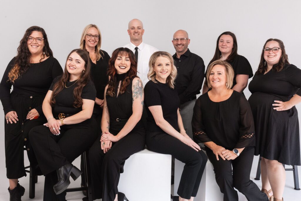 Ten people pose together in a studio against a white background, all dressed in black attire. Some are seated on stools while others stand behind them, smiling at the cameraâ€”capturing the spirit of a close-knit group from Greeley.
