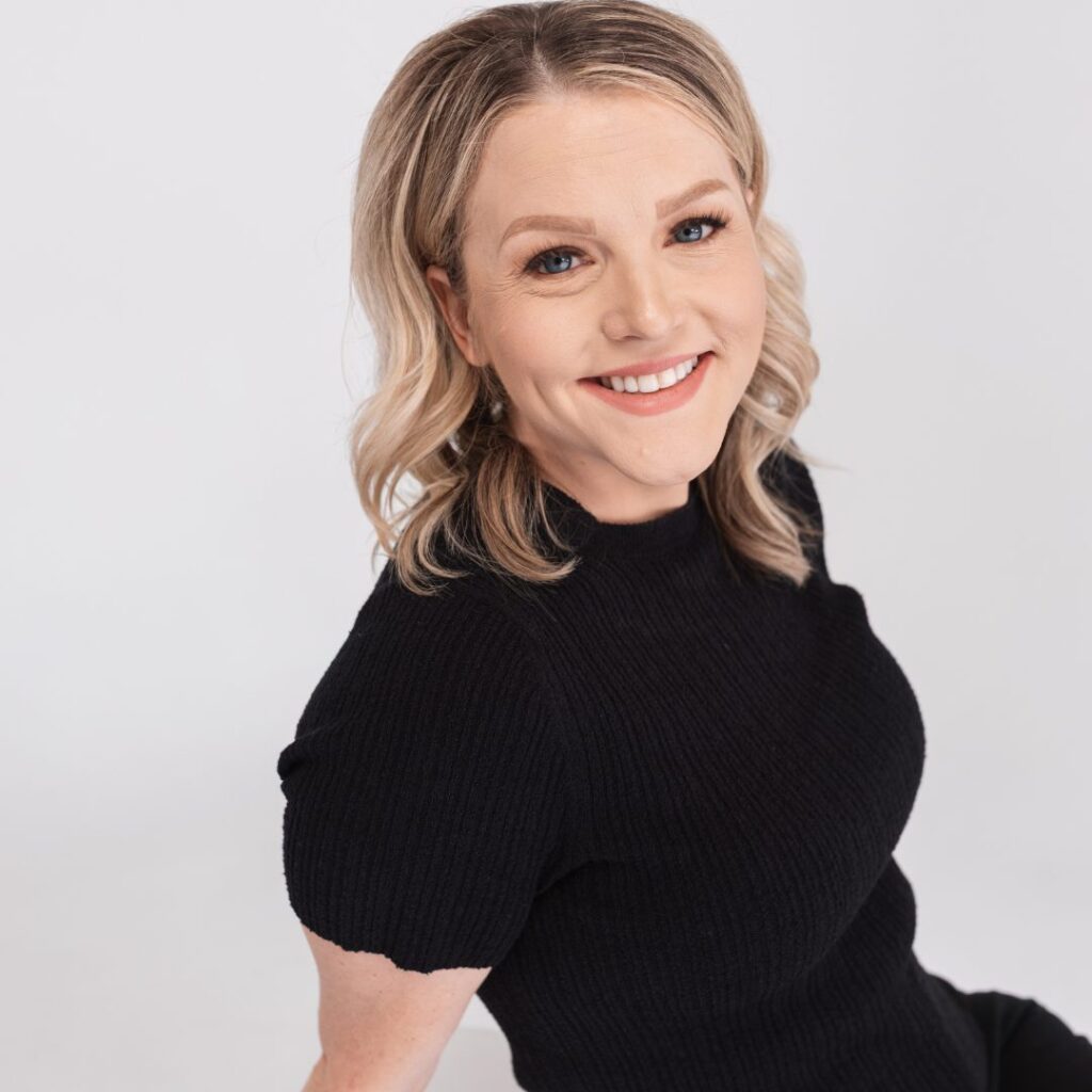 A woman with shoulder-length blonde hair and light skin is smiling at the camera. She is wearing a short-sleeved, black ribbed top and posed against a plain white background, radiating warmth and approachabilityâ€”perfect for any team setting.