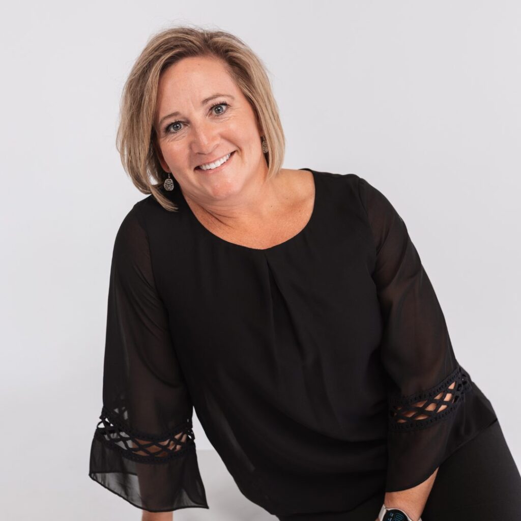 A woman with short blonde hair, wearing a black blouse with lace sleeves, smiles at the camera against a plain white background, radiating confidence and warmthâ€”perfect for any team introduction.