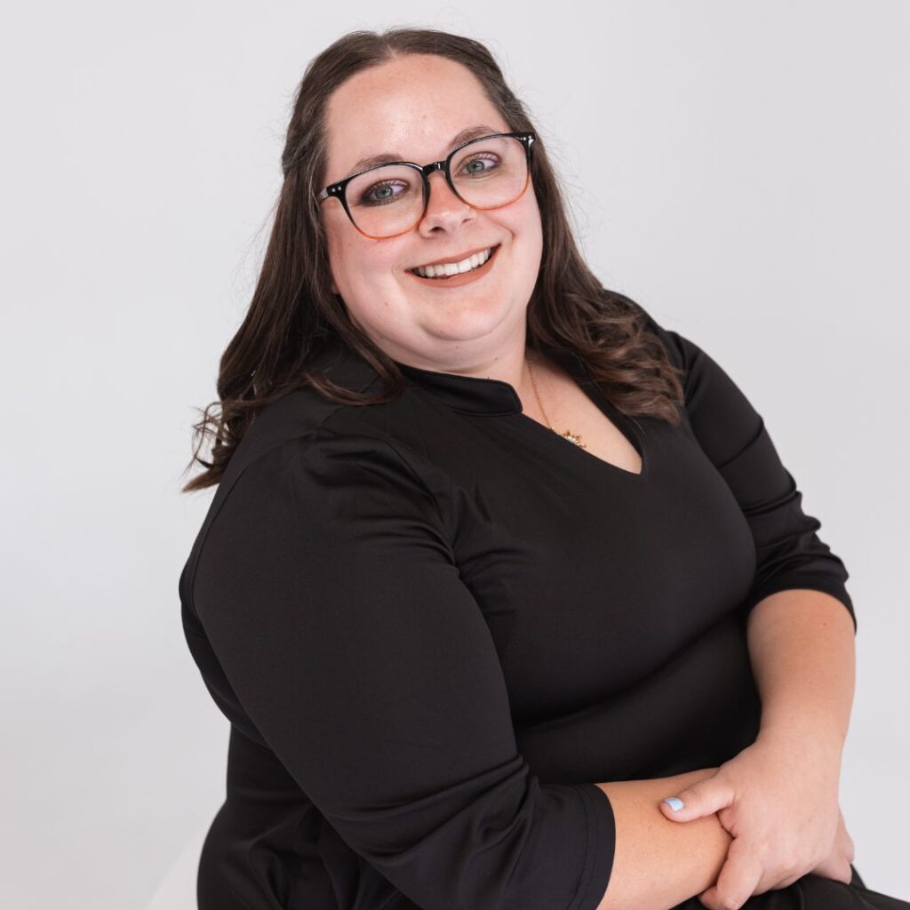 A woman with long brown hair, wearing glasses and a black dress, smiles while sitting with her hands folded on her lap against a plain white background, ready to join your team.