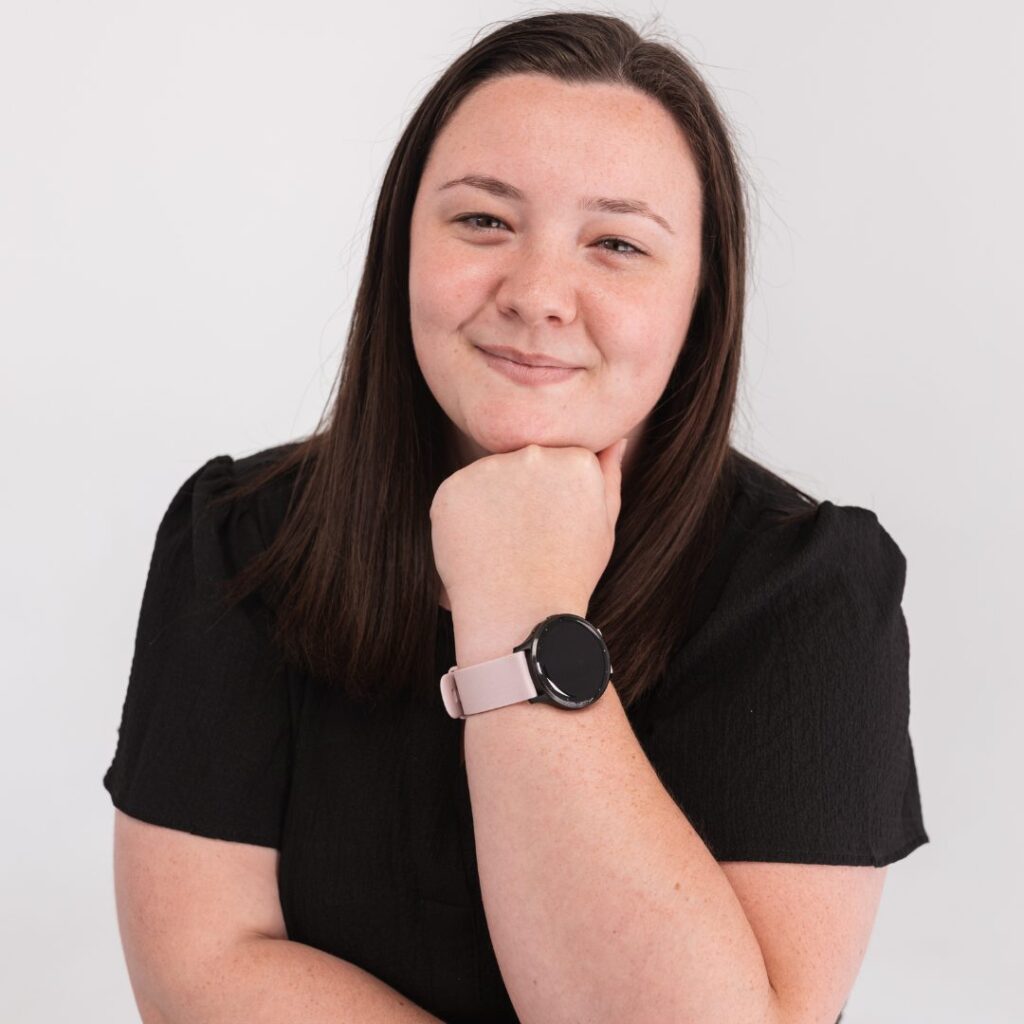 A woman with straight brown hair, wearing a black top and a smart watch with a light pink band, smiles softly while resting her chin on her hand, exuding calm confidence as a valued team member against a plain white background.