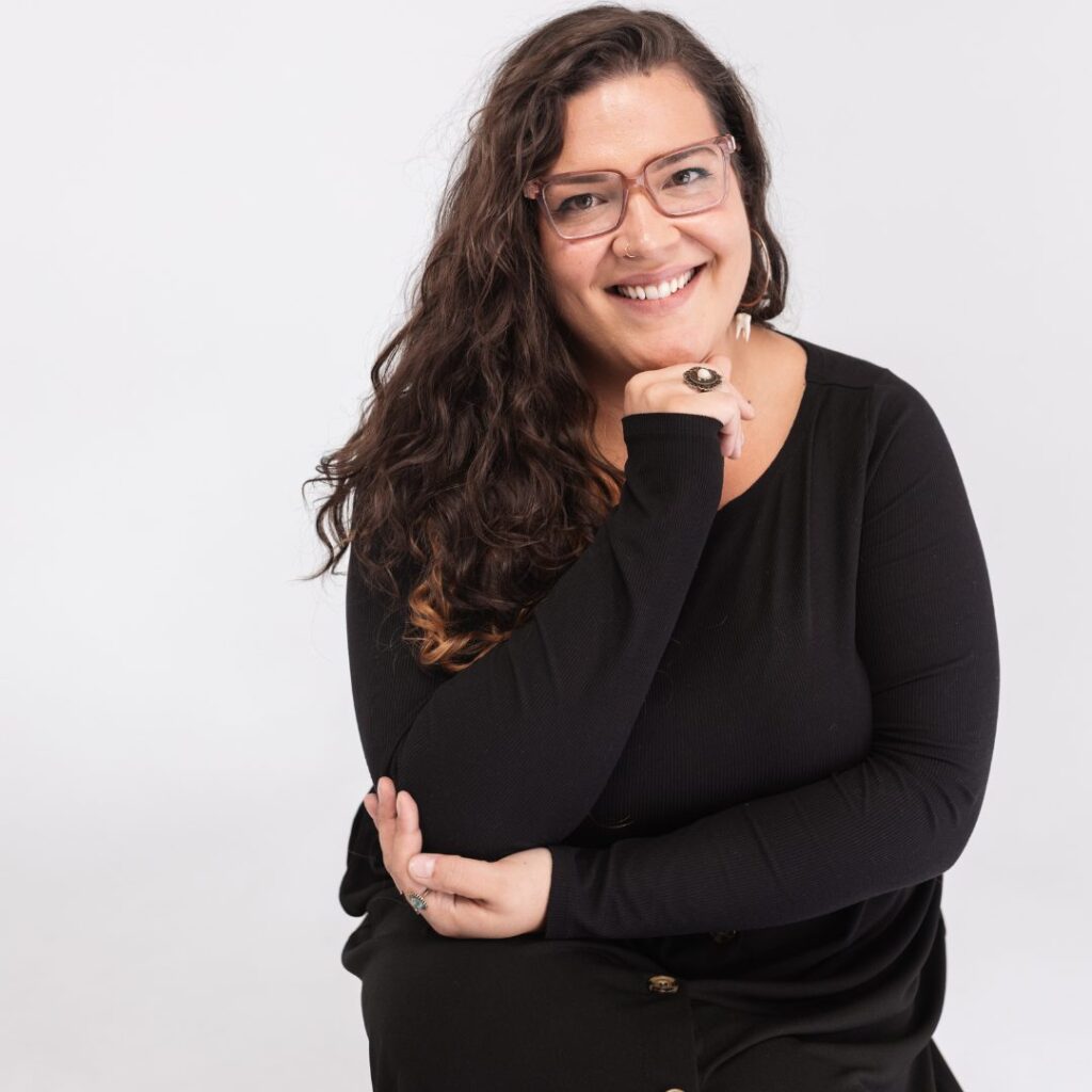 A woman with long, wavy brown hair and glasses, wearing a black top, sits smiling with her hand resting by her chin against a plain white background, embodying confidence and approachabilityâ€”perfect for any team setting.