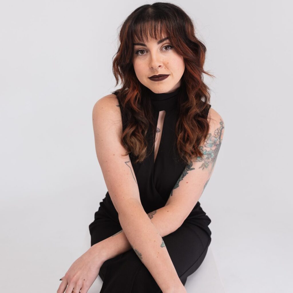 A woman with wavy brown hair, dark lipstick, and floral tattoos on her arm sits on a white chair against a plain white background, wearing a sleeveless black outfit and looking confidently at the camera, embodying strong team spirit.