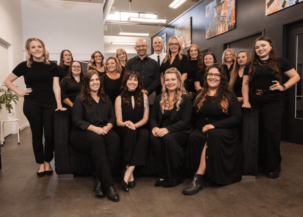 A group of nineteen people, mostly women and a few men, dressed in black outfits, pose and smile together indoors in the modern office space of Northern Colorado Periodontics. Some are seated on a couch while others stand behind them.