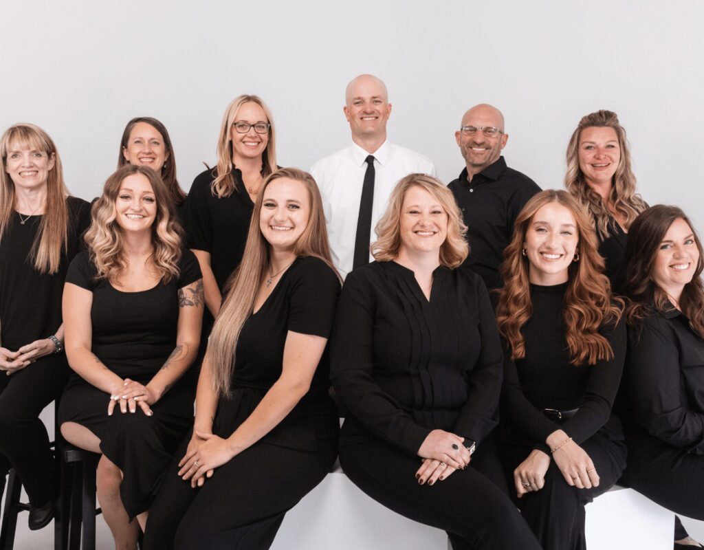 A group of twelve people, mostly women and one man, pose together in coordinating black outfits against a plain light background. Smiling warmly, they appear to be part of a professional Fort Collins team or office staff.