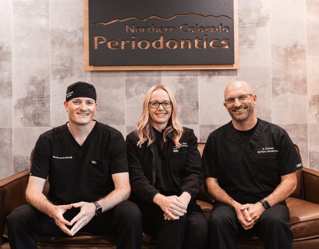 Three smiling periodontists in black uniforms sit on a brown couch beneath a sign that reads Northern Colorado Periodontics in an office setting.