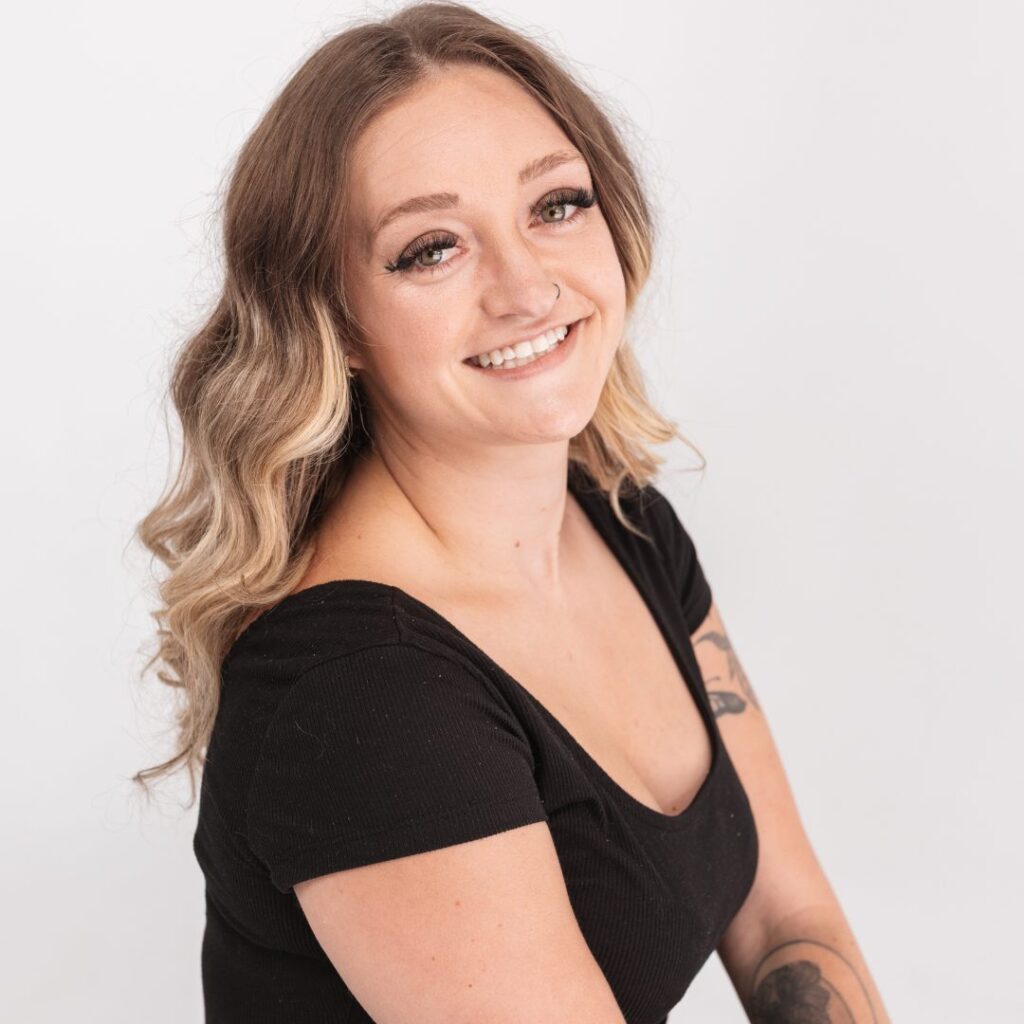 A smiling woman with wavy, light brown hair and a nose ring, wearing a black top, sits against a plain white background. She has a visible tattoo on her upper arm and exudes a friendly, team-oriented vibe.