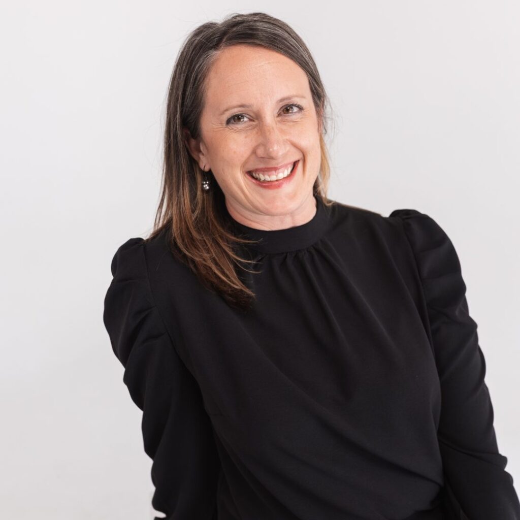 A woman with straight brown hair and a friendly smile is wearing a long-sleeved black top. She sits against a plain, light background, radiating the approachable energy of a valued team member.