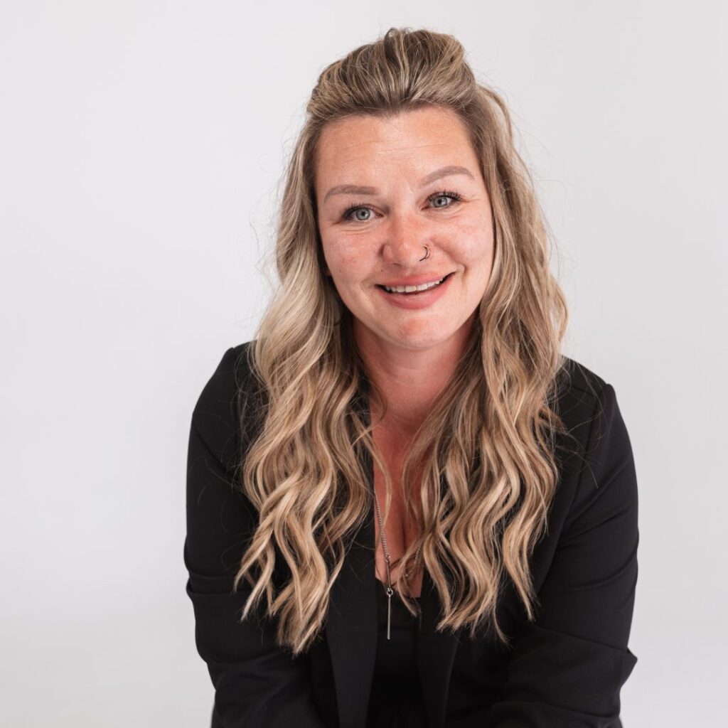 A woman with long, wavy blonde hair and a nose piercing smiles at the camera. She is wearing a black top and blazer, seated against a plain light background, ready to join the team with her approachable and confident presence.