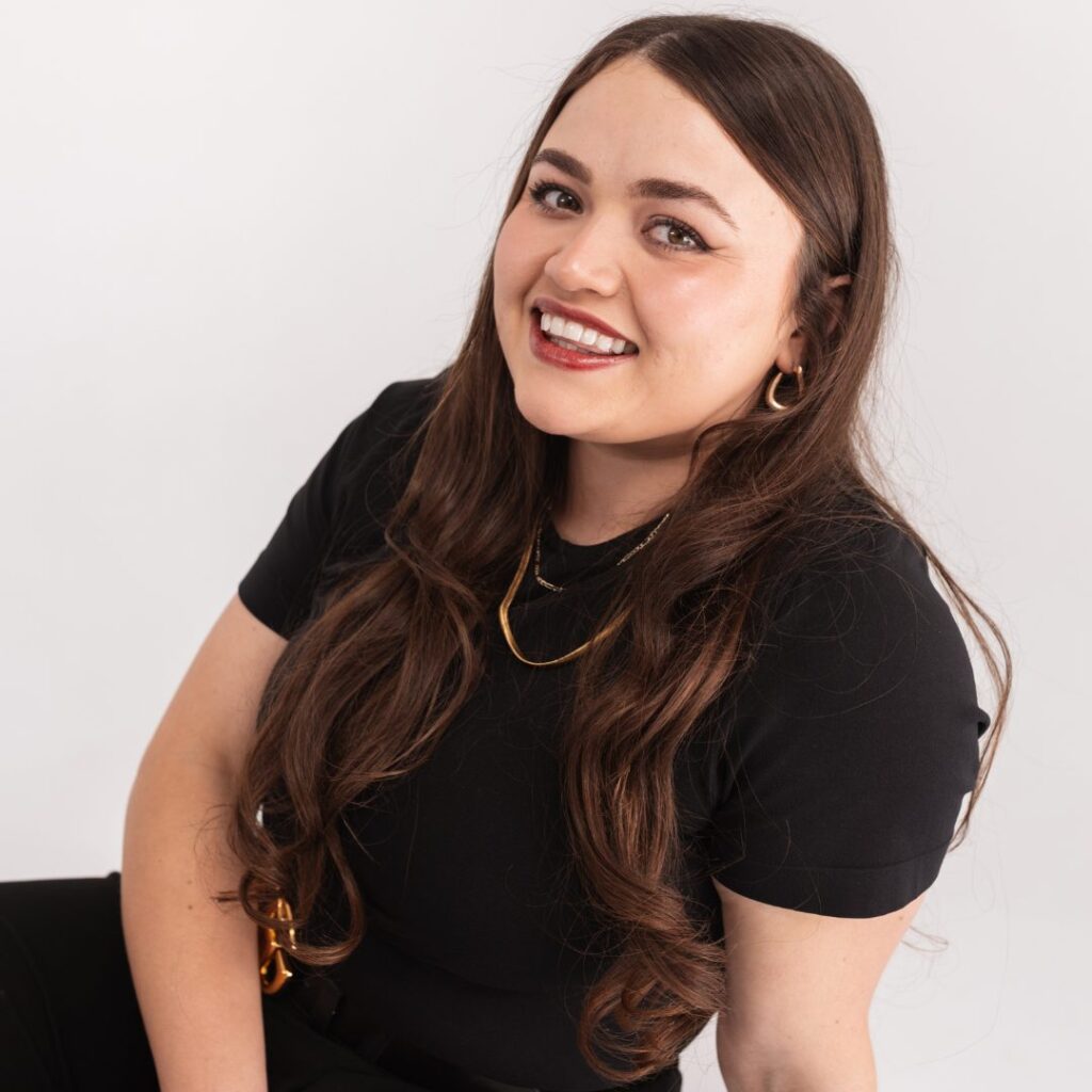 A young woman with long brown hair, wearing a black top, gold necklace, and hoop earrings, smiles at the camera against a plain white background, ready to join the team.