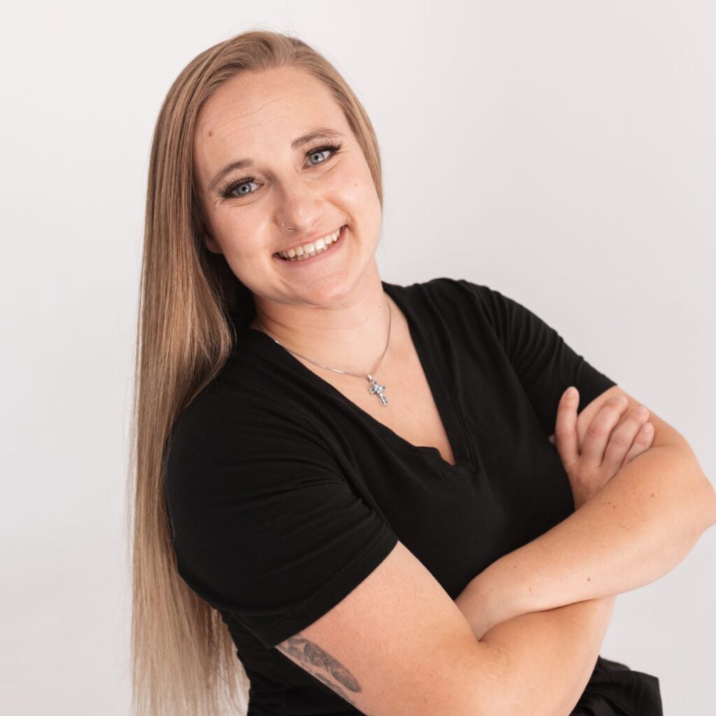 A woman with long straight blonde hair wearing a black shirt stands with her arms crossed, smiling confidently at the camera against a plain white background. She has a tattoo on her forearm, wears a necklace, and exudes team spirit.