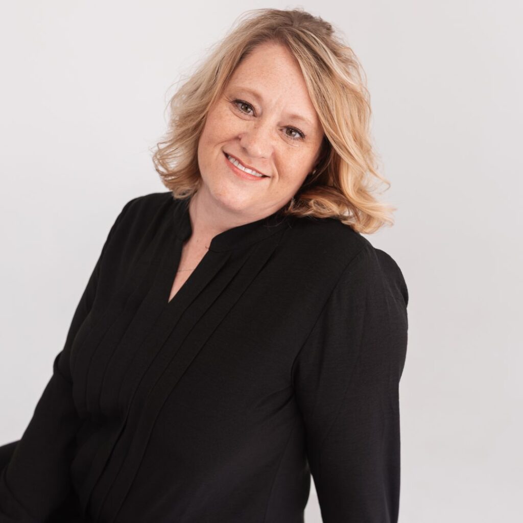 A woman with blonde, wavy hair and light skin smiles at the camera. She is wearing a long-sleeved black blouse and poses confidently against a plain white background, radiating warmth and approachabilityâ€”an ideal addition to any team.