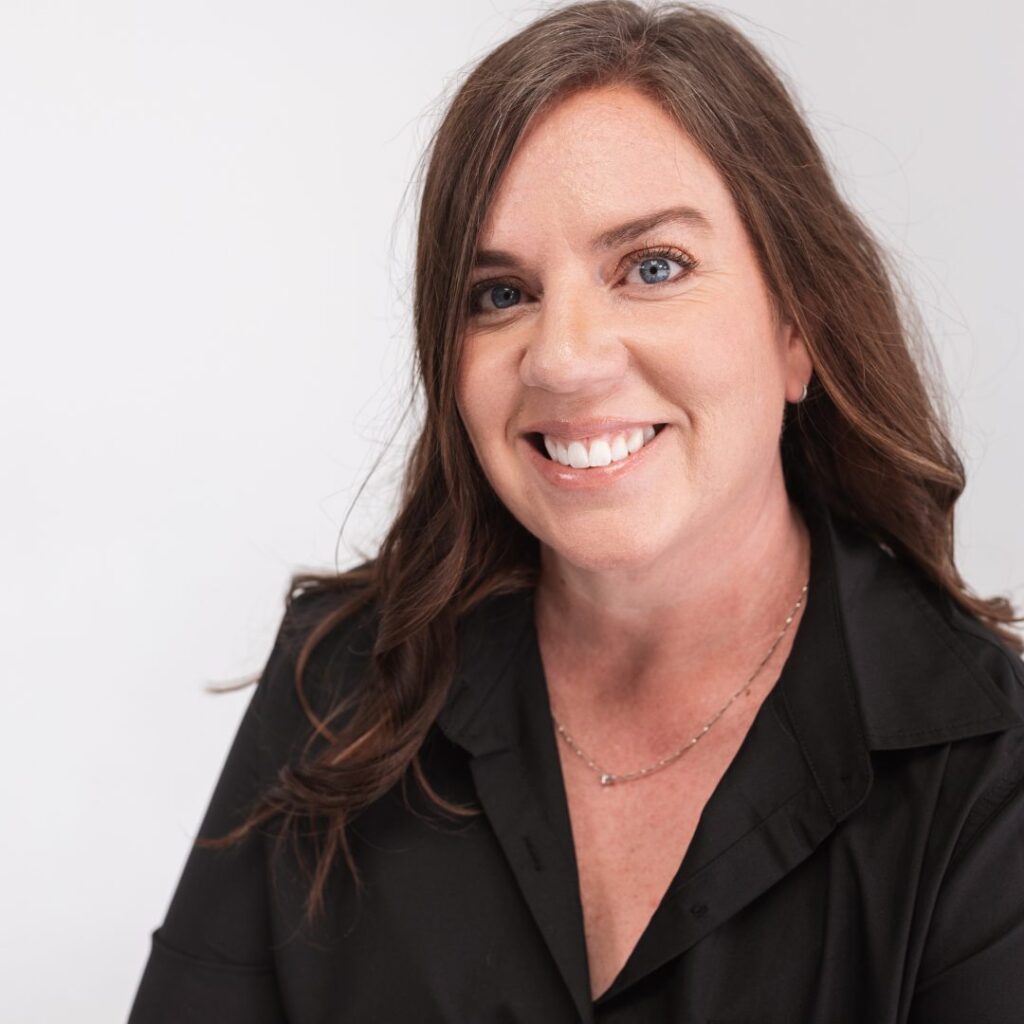 A woman with long brown hair and blue eyes smiles at the camera. She is wearing a black blouse and a delicate necklace, posed against a plain white background, radiating confidence as a valued member of her team.